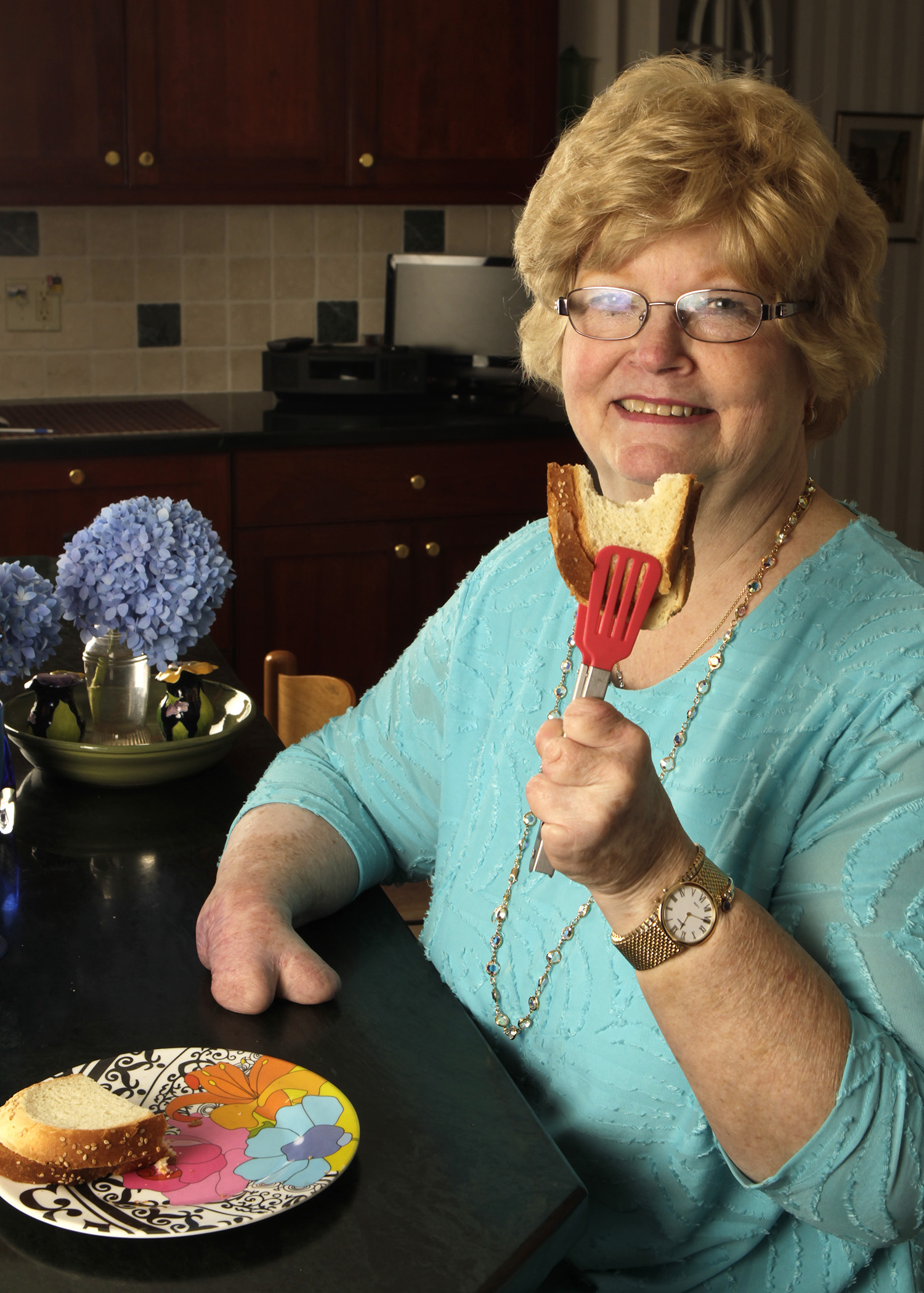 Image of Cindy sitting at a table, holding up a sandwich in red-silicone-tipped metal tongs, smiling at the camera.
