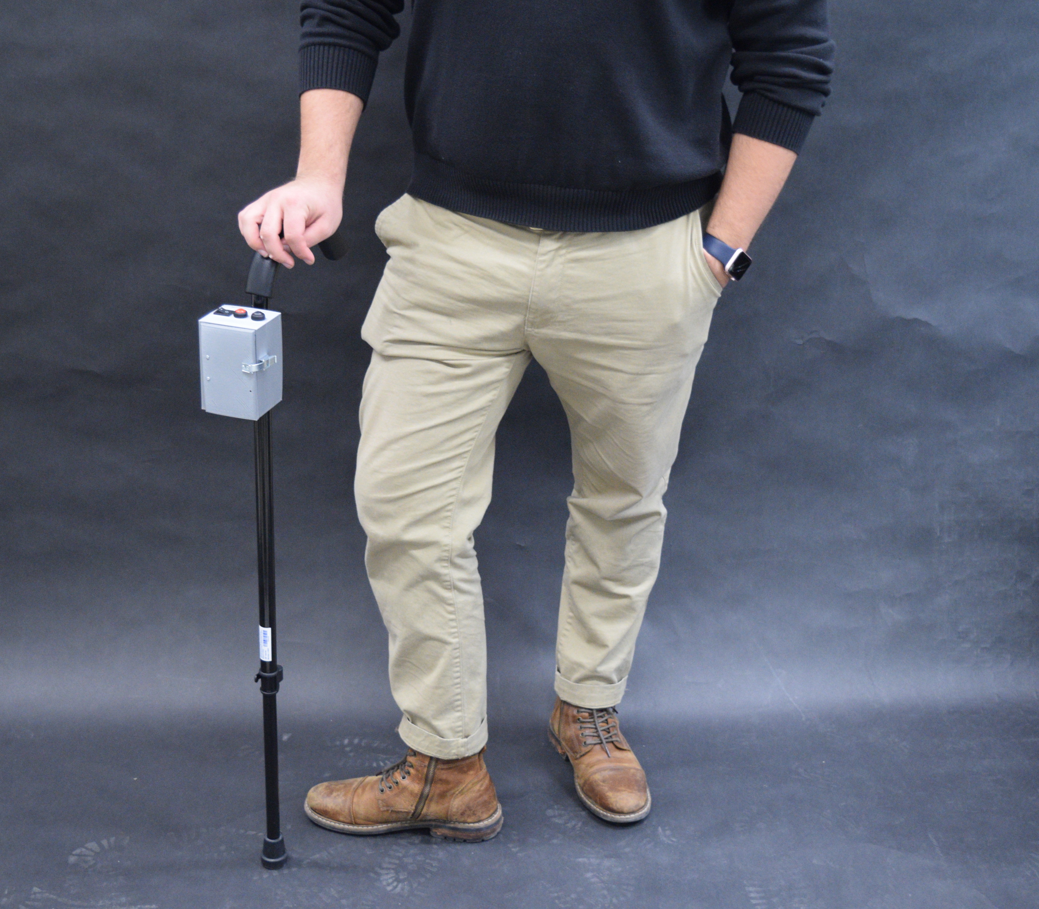 Photograph of a person posing with a cane; the cane has a medium-sized grey plastic box affixed near the top.