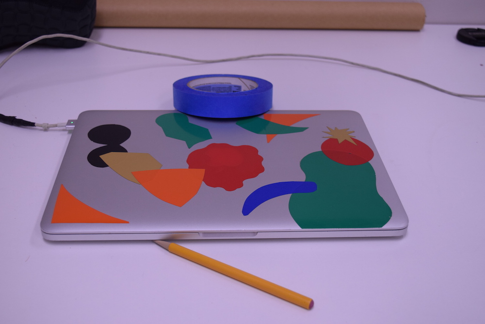 Image of silver laptop with colored stickers on it, sitting on a white table, with a yellow pencil and blue roll of tape. The whole image has a purple cast to it.