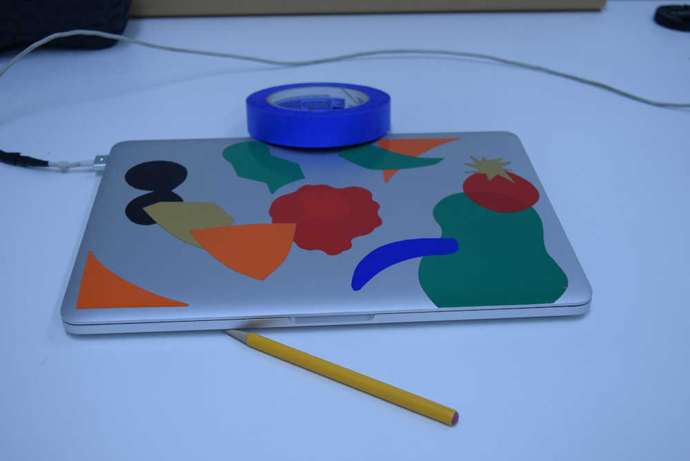 Image of silver laptop with colored stickers on it, sitting on a white table, with a yellow pencil and blue roll of tape. The whole image has a distinctly blue cast to it.