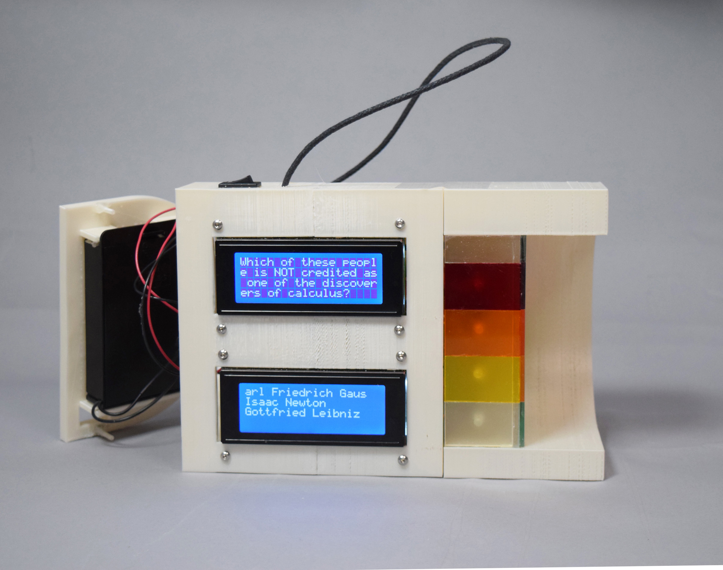 Photograph of a rounded white plastic device with an open side compartment showing a battery holder, a front panel with two large text display screens, and five colored stripes along the right edge.