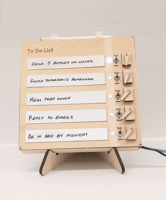 Brief gif of a wooden box with handwritten white fields with text like "finish tomorrow's homework," "reply to emails," etc. Each message has a lit white LED and toggle switch next to it, and a user flips the switch down to mark each task as done. When she's finished, each toggle switch gets returned to the upwards position by a small motor with a wooden arm.