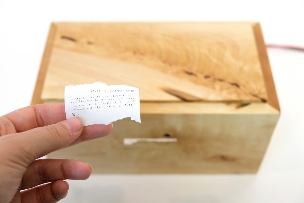 An image of a hand holding a small printed receipt with mostly illegible paragraph-form text written on it. The first line of the text is a date and time. In the background, a closed wooden box with beautiful grain. A small piece of receipt paper is torn and emerging from the front of the box, suggesting that the piece being held in the foreground was torn from that source.