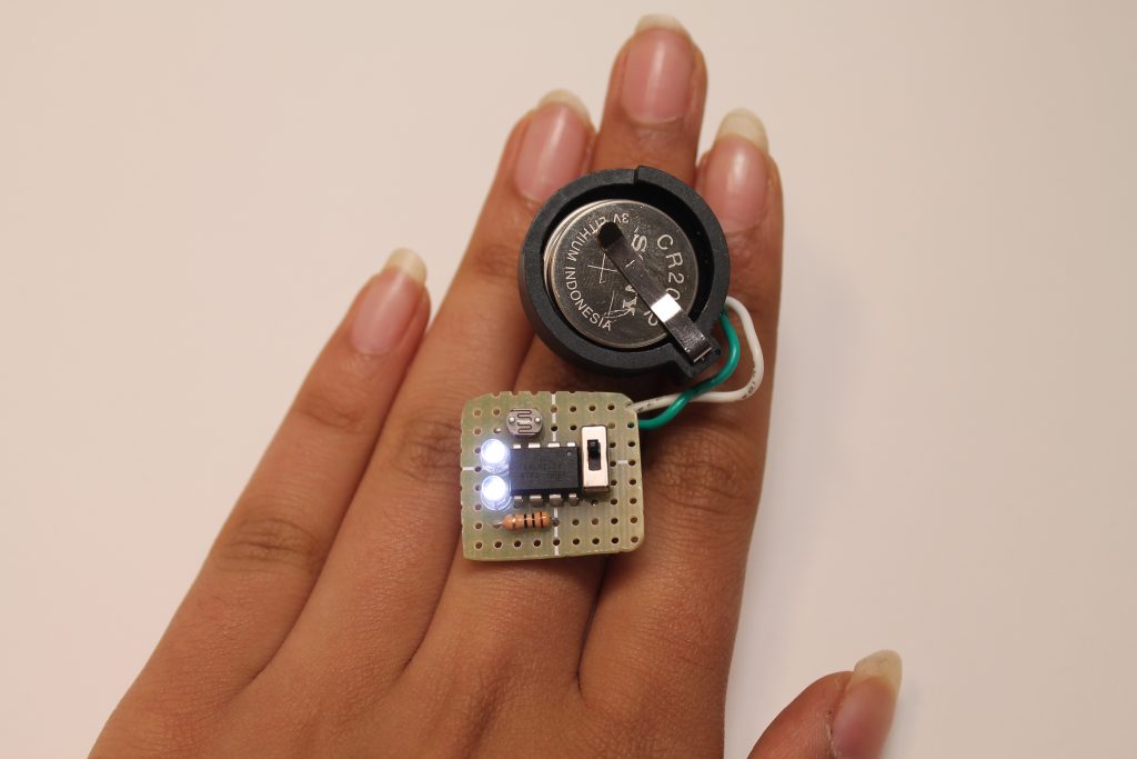 Image of two unusual rings on a person's left middle finger. One ring is a battery holder for a large coin-cell battery. It is connected by a short length of green and white wires to the other ring, which is a small square section of perforated circuit board containing two illuminated LEDs, a photoresistor, a resistor, an 8-pin IC, and a sliding switch.