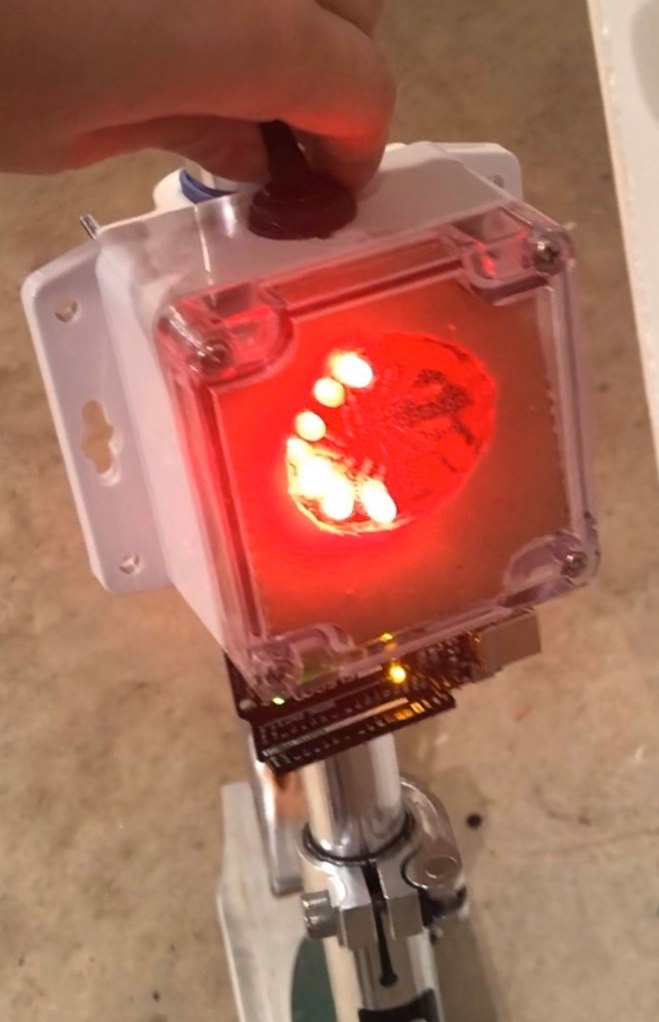 Photograph of a clear-faced plastic box affixed to a scooter's vertical pole; the top of the box has a toggle switch pointed to the left, and through the front of the box, a half circle of red LEDs are illuminated to the left.