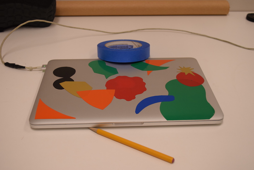 Image of silver laptop with colored stickers on it, sitting on a white table, with a yellow pencil and blue roll of tape. The whole image has a weak pink cast to it.