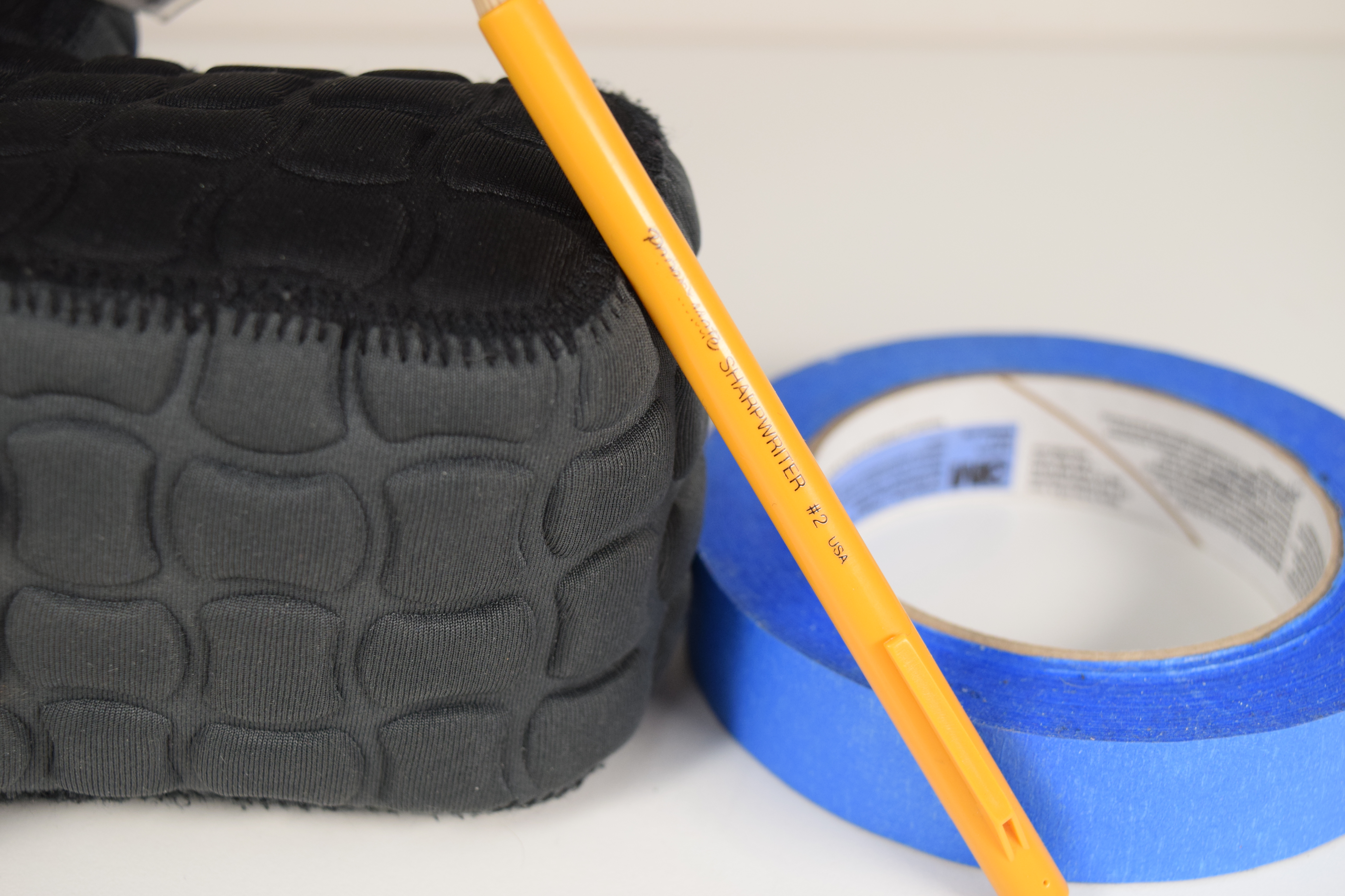 Still life of a yellow pencil leaning against a grey and black camera case, with a blue roll of masking tape behind it.