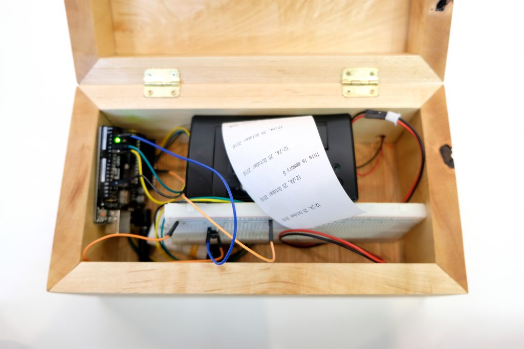 Image of an opened wooden box. Inside of it is a thermal receipt printer with paper coming out of it and a string of short printed messages with gaps between them.