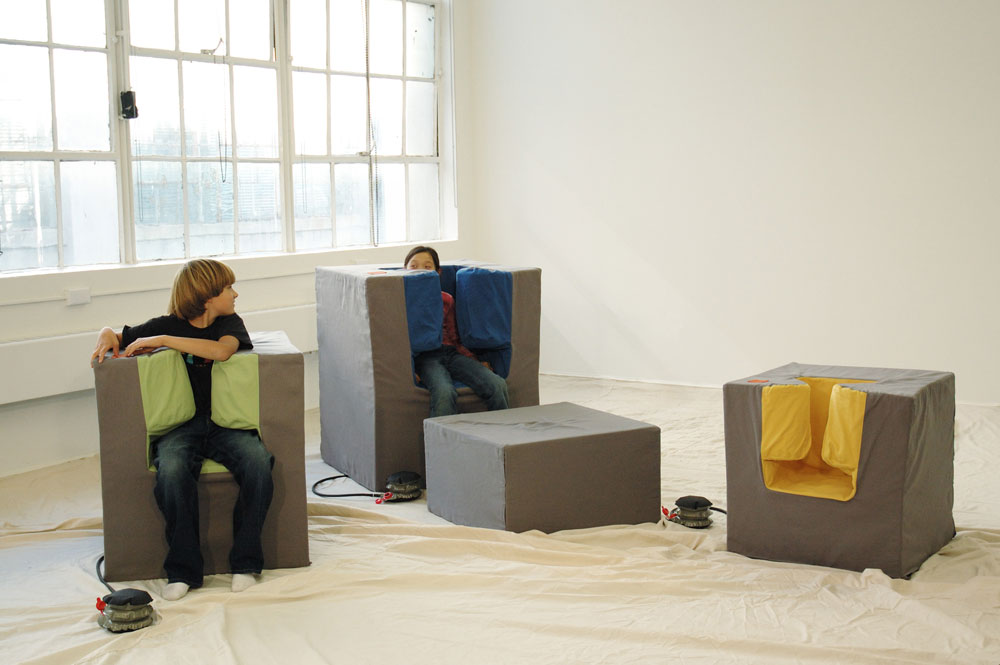 Image of three chairs in a room. The leftmost chair has a young boy sitting in it, and he is nearly surrounded by an inflatable material which is hugging him. The center chair has a young girl in it; the inflatable material nearly covers her head up and she peeks out to the side.