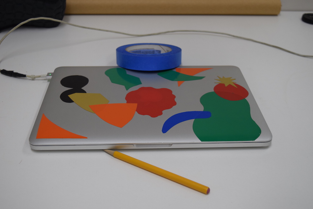 Image of silver laptop with colored stickers on it, sitting on a white table, with a yellow pencil and blue roll of tape. The whole image has no discernible color cast to it and the whites appear white.
