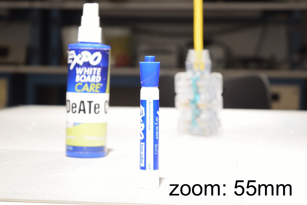 Sequence of five images with zoom moving from 55mm to 18mm sequentially. A marker is in the foreground and in the background are a bottle to the left, then farther back to the right a power cord, and then finally the wall of the room. As the zoom moves from 55mm to 18mm, the marker remains roughly the same size while the background objects become relatively smaller.