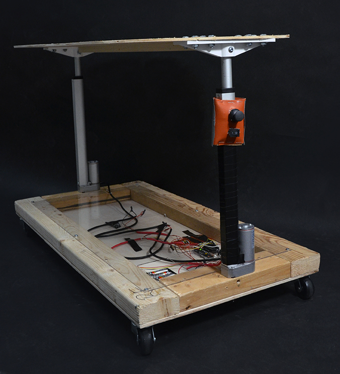 Photograph from the edge of a wooden elevator device, with a wheeled rectangular base and two telescoping metal arms supporting opposite sides of a wooden top.