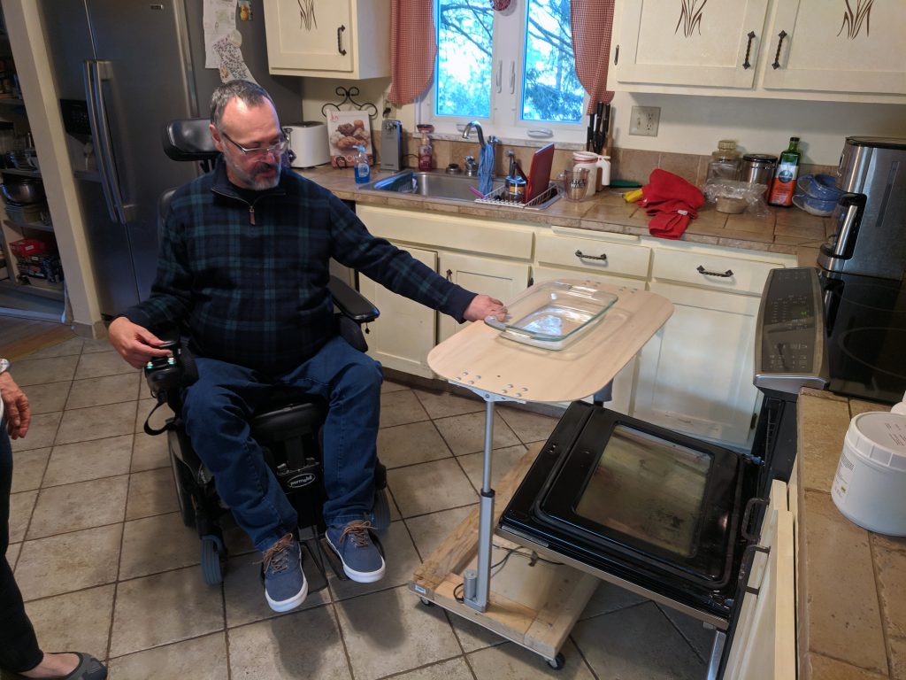Photgraph of a man in a wheelchair in his kitchen reaching out to an elevated wooden tray with a glass baking tray on it. The wooden tray is suspending the tray over an open oven door.