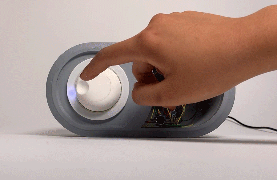 Moving image of a hand turning a white knob on a medium-sized rounded grey box; as the knob is rotated, a blue light ring around it changes its illumination to follow the turning.