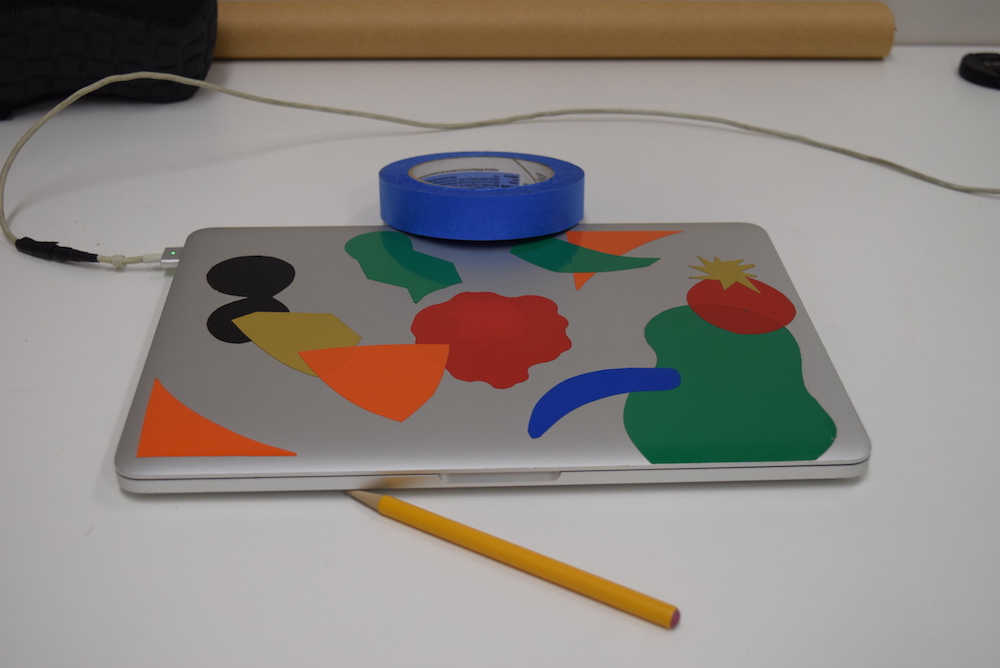 Image of silver laptop with colored stickers on it, sitting on a white table, with a yellow pencil and blue roll of tape. The whole image has no discernible color cast to it and the whites appear white.