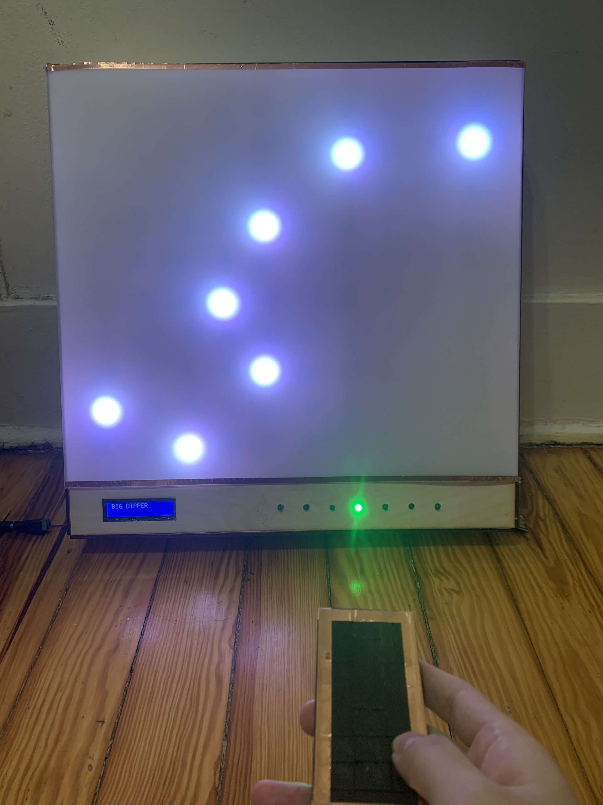 Photograph of a white translucent sheet of paper above a wooden-framed box with an LCD display and some illuminated green LEDs. Behind the sheet are seven white circles of light arranged like the Big Dipper constellation. In the foreground, a hand holds a small black-covered remote control.