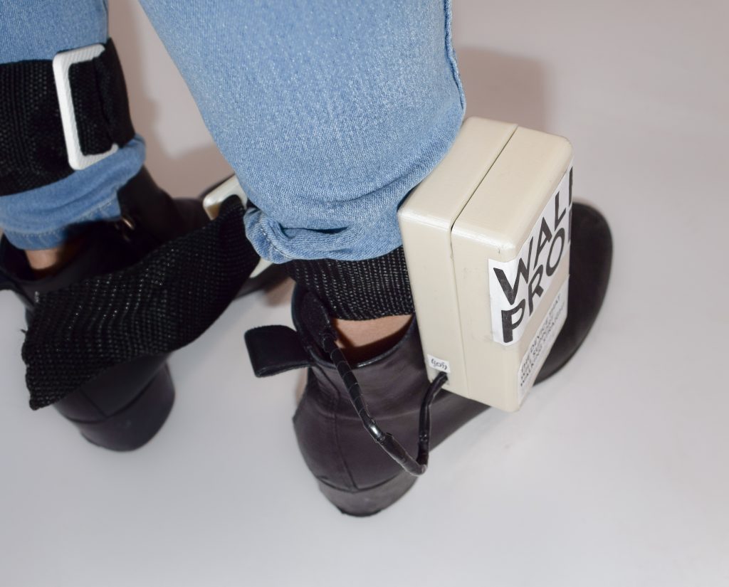 Photograph of a beige plastic box strapped to a person's right ankle, with the words "Walk Prop" labeled on the side.