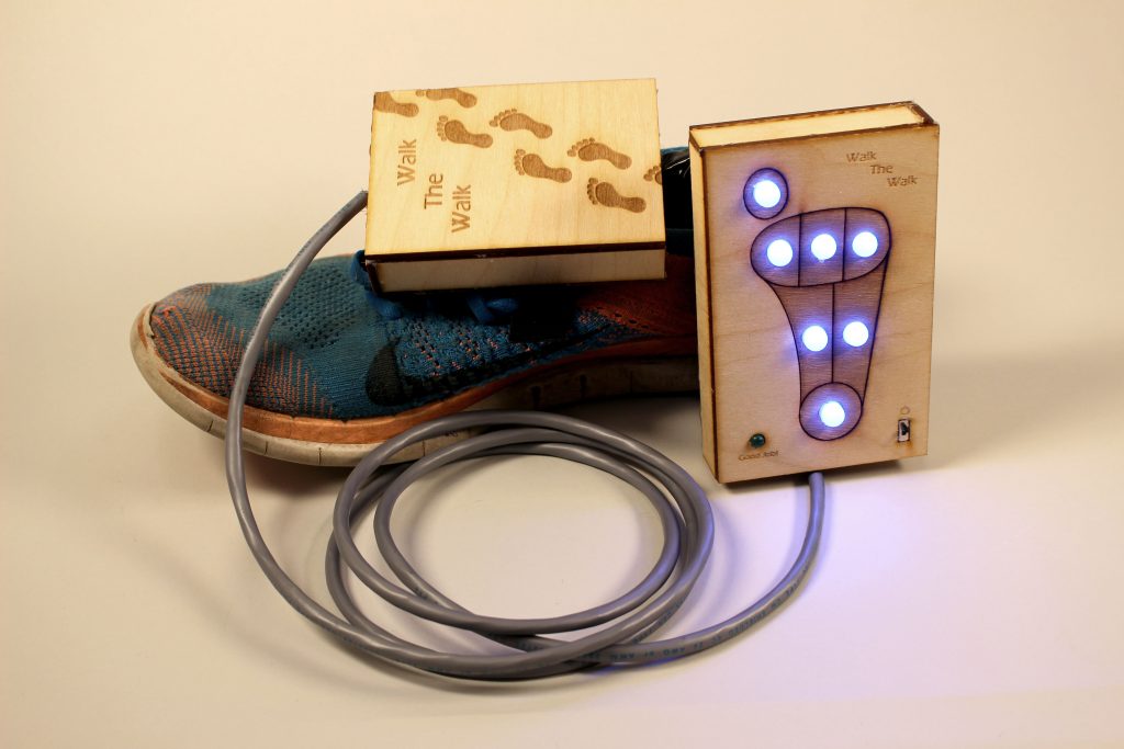 Image of a shoe with two lasercut wooden boxes nearby, connected by cables. One box is labeled "Walk The Walk", and the other one has a simple diagram of a foot on its face, with seven LEDs located at particularly defined pressure points.