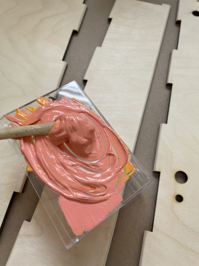 There is a blob of coral-pink paint mixed on top of a clear acylic lid. Behind the paint are blank planks of plywood