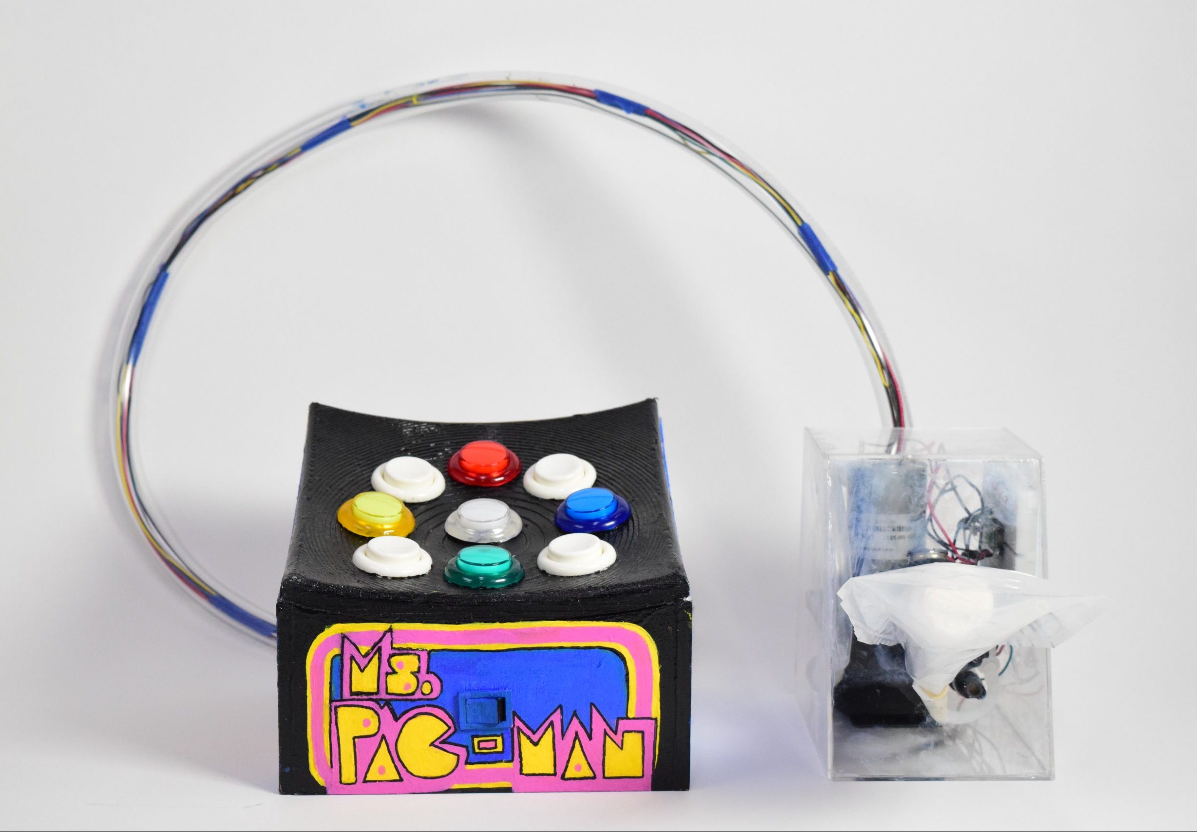 This picture shows a black box with a curved surface. There are 9 colored arcade buttons on its surface. On the front side of the box there "Ms. Pac - Man" is drawn in graphic letters. There is a clear tuber with wires inside that connects the black box to an acrylic casing that holds more wires. There is a joy stick attached to the acrylic casing.