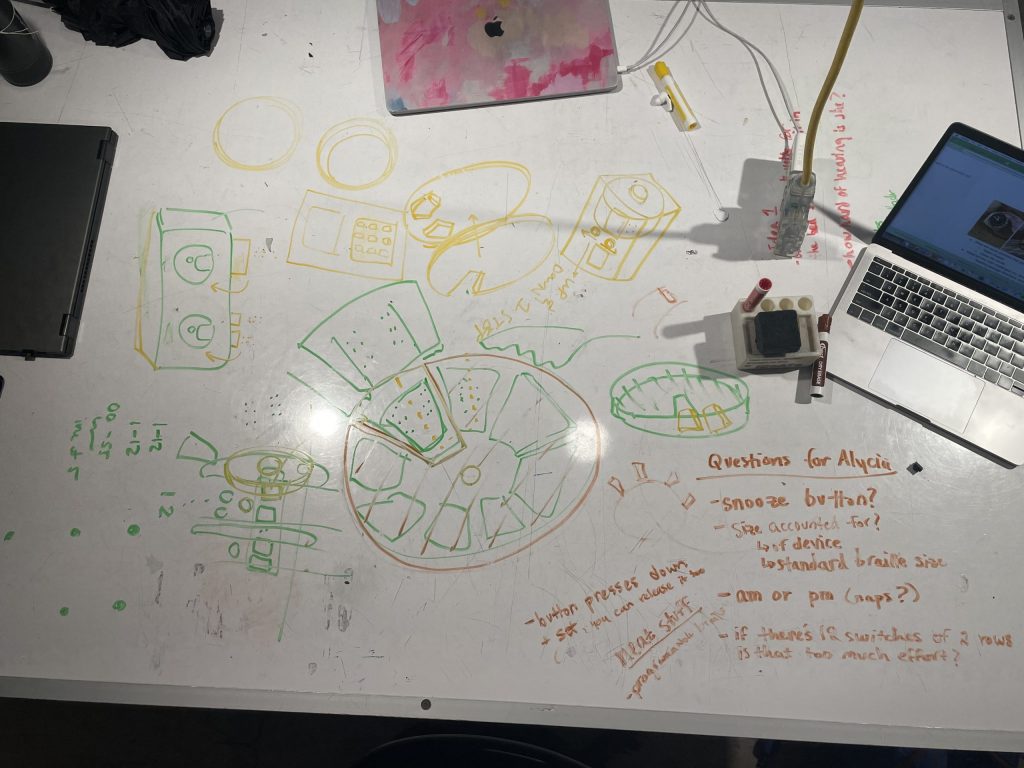On a whiteboard table are drawings in yellow, green, and red, depicting different ideas for our clock