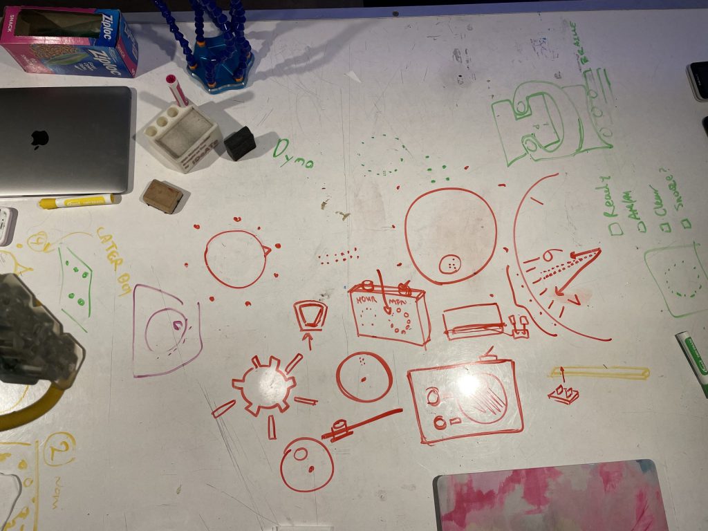 In the center of the image are red marker drawings depicting different clock ideas, around it are 2 closed laptops and 3 markers strewn on the table