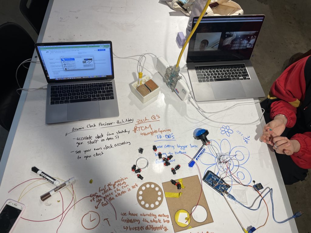 There are 2 laptops open on a table and in front of them are marker drawings and writings about different clock ideas. Some buttons, wires, and chipboard are also next to the writings