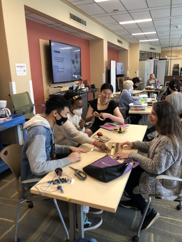 Seated around a table are 4 people. On the right is Alycia, a middle-aged woman with long hair. On the left are 3 students, David, Noni, and Catherine who are handing Alycia chipboard cutouts to try as clock interfaces
