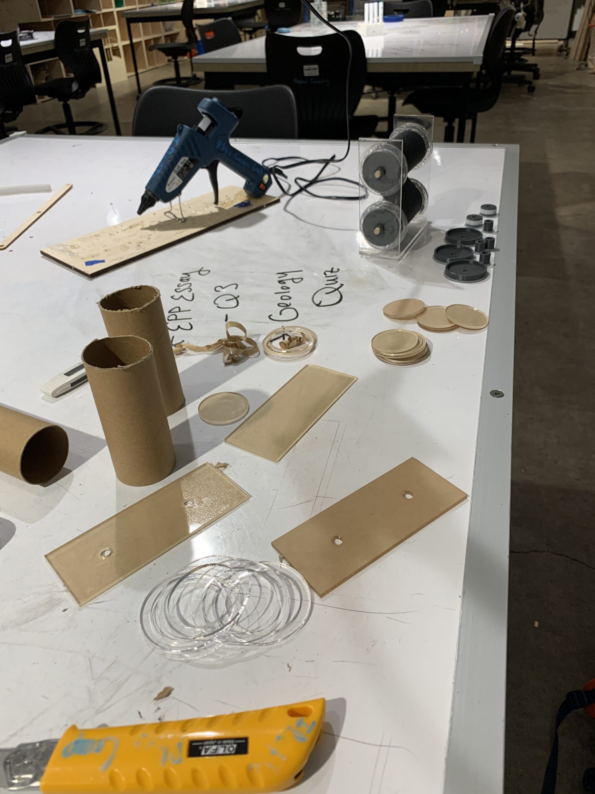 a photograph of a table with various parts for constructing the bill rollers. There are a couple of toilet paper tubes, rectangular pieces of acrylic with two holes in each, a hot glue gun, and a couple clear acrylic rings. 