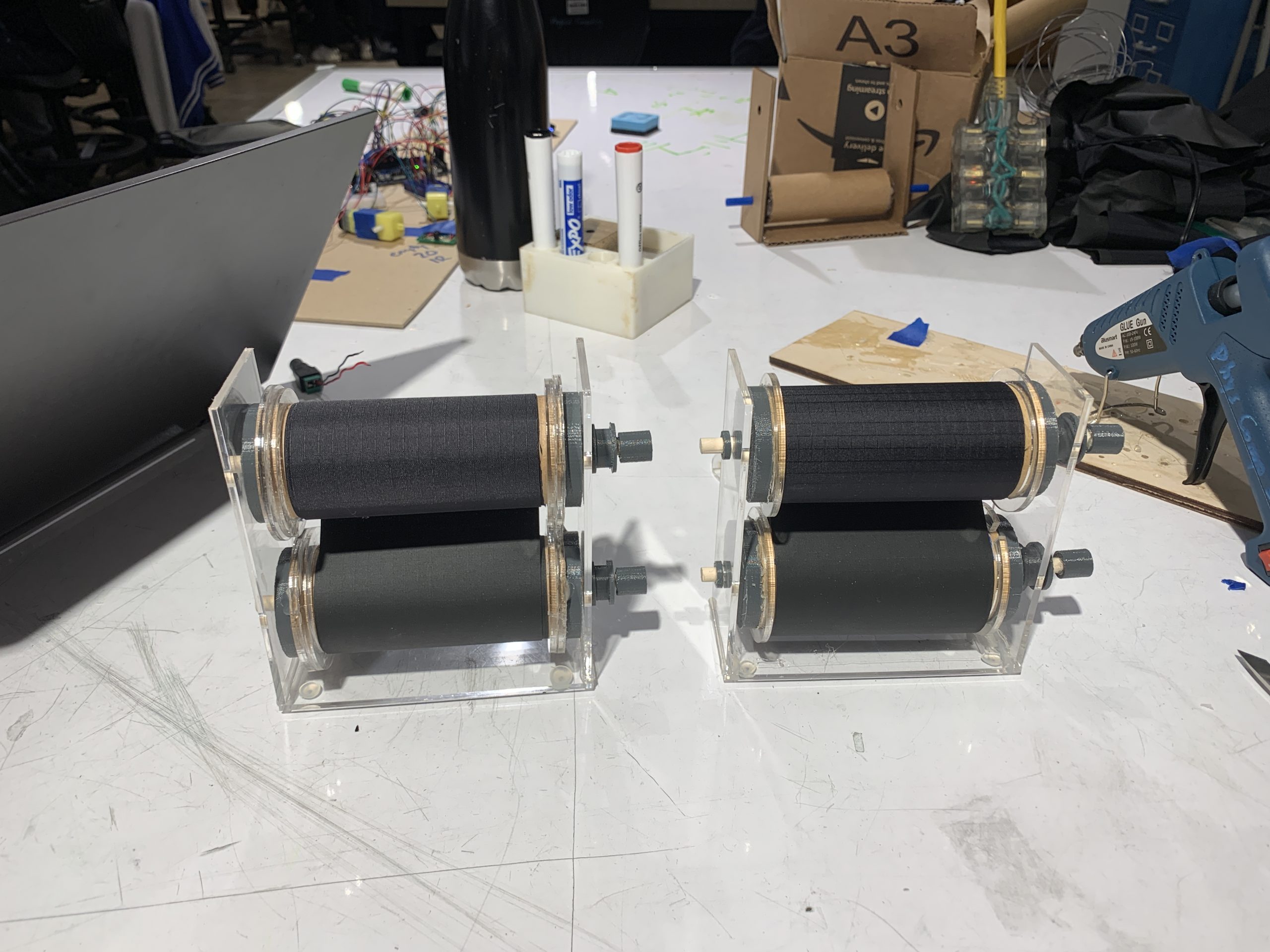 Two fully-constructed bill rollers. These rollers are made from toilet paper tubes wrapped with black nylon fabric, and have gray 3D-printed connectors on the end to pair with the toy gear box motors.