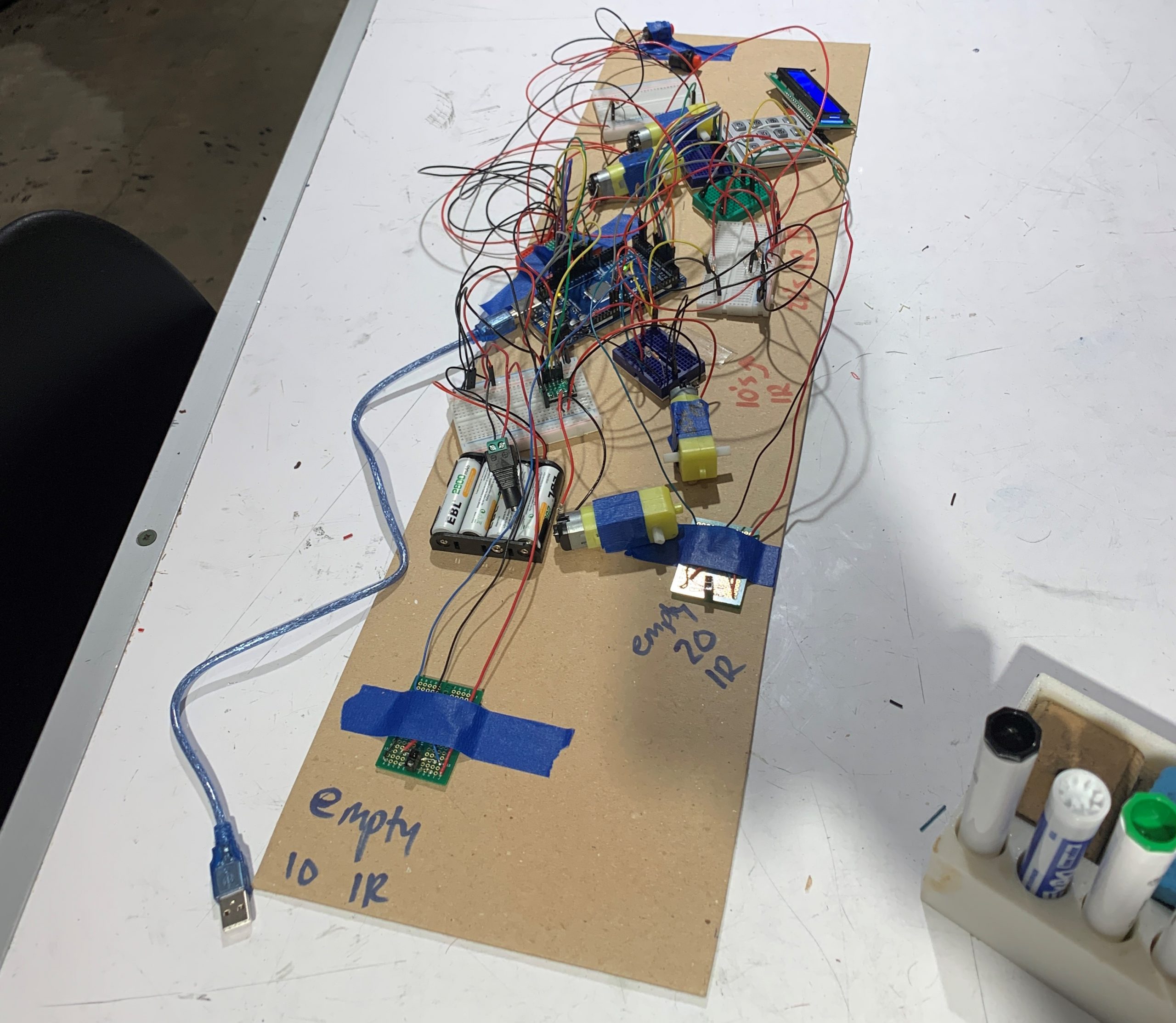 This image includes our hardware testing set-up. There is a long piece of cardboard with many electrical components and wires such as motors, a keypad, a microcontroller, and IR sensors.