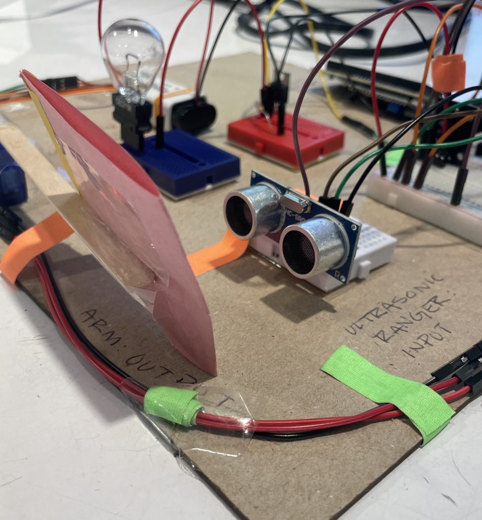Image of a breadboard holding an ultrasonic ranger that is positioned in front of a piece of paper attached to a servo motor arm made of a popsicle stick.