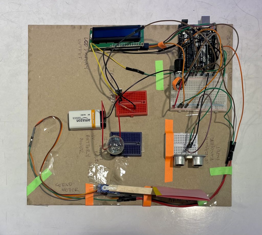 Image of an arduino set-up taped to a piece of chipboard. The set-up starts by connecting the arduino to a breadboard that has multiple wires and a rotary encoder plugged in. The wires connect a LCD display, servo motor, ultrasonic ranger, transistor, and battery to the breadboard. The transistor is also connected to an incandescent bulb.