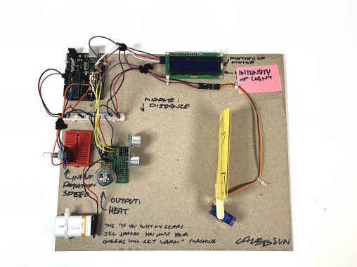 A roughly A4 sized piece of chipboard with Arduino components on it. There is a small LCD screen, a servo motor with a piece of yellow paper attached, as well as a few mini breadboards and a 9 volt battery. The wires are organized with pipe cleaners and the pieces are held to the chipboard with zip ties