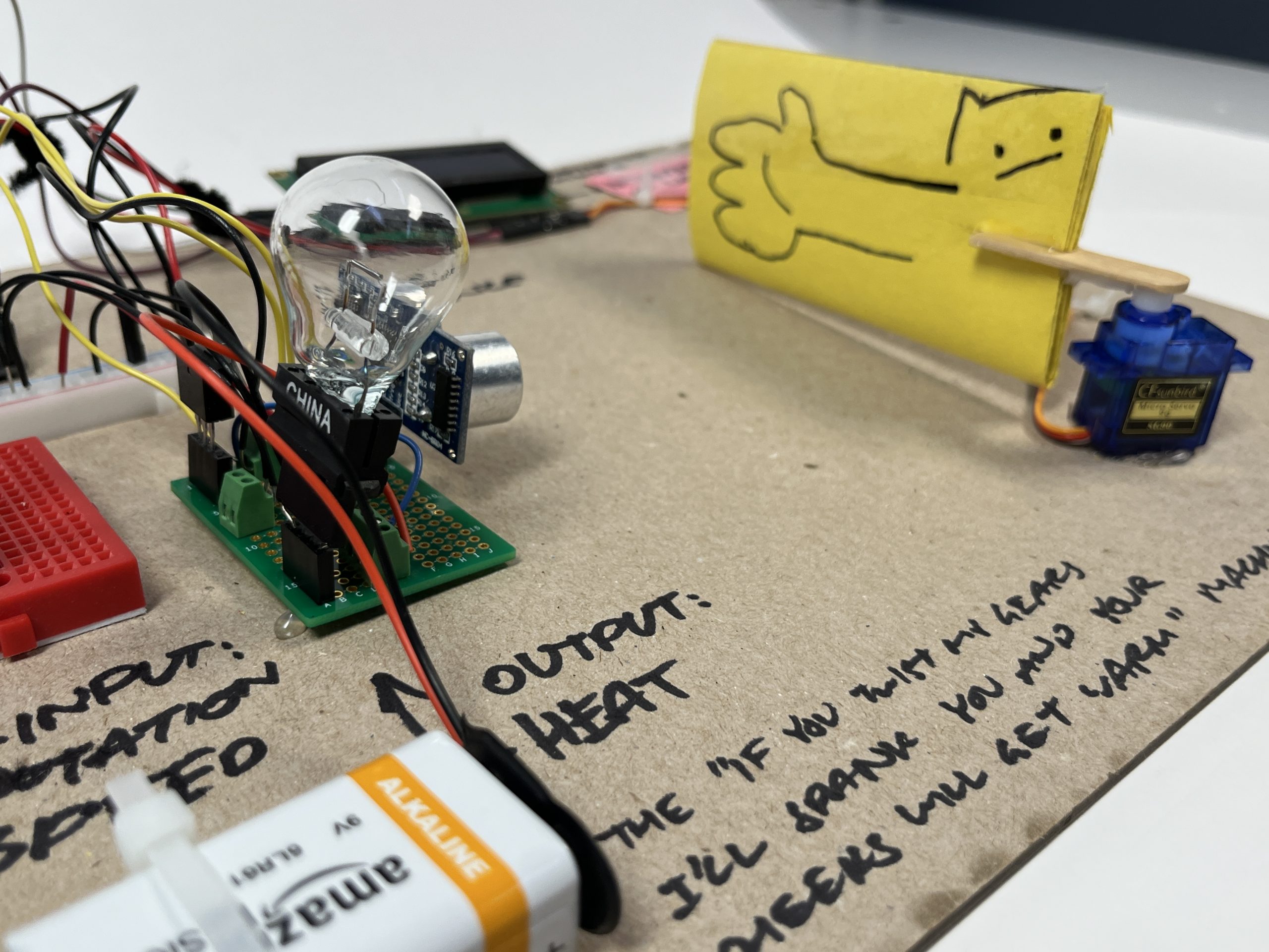 A closeup shot of the transducer, with emphasis on the yellow paper arm that is attached to the servo motor. It has a big cartoonish hand drawn on it with a cat in the background.