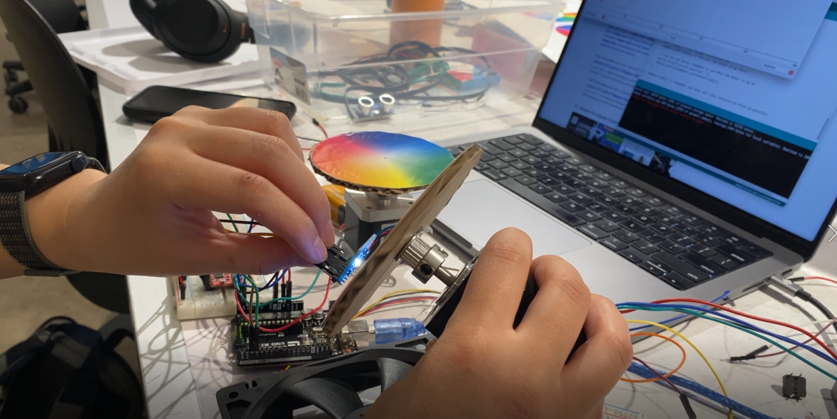 A hand holding the adafruit sensor next to a color wheel, propped up on a stepper motor.