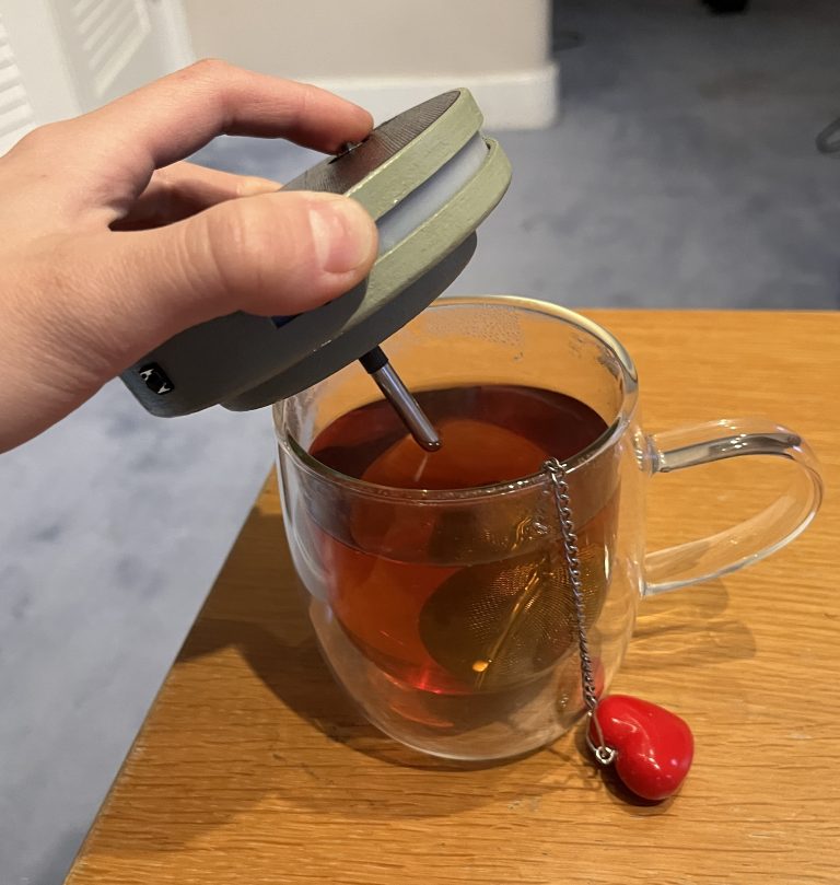 A person holds an olive green 3d printed plastic disc in his left hand; the disc is 5" wide and 1" thick. Sticking out of the bottom of the disc is a narrow shiny metal probe. The entire disc and probe assembly is held above a clear mug of hot tea.