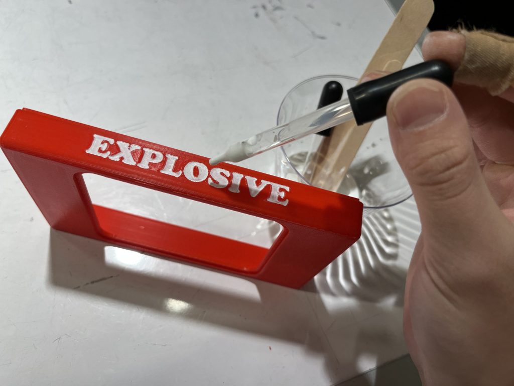 Half of a red 3D-printed box standing on its side with "Explosive" written in white. A hand holding a dropper with white paint inside is reaching towards the letters