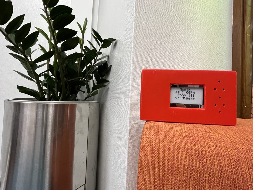 Red box with a small screen in the center and 7 holes to the right side, next to a plant and on an orange surface