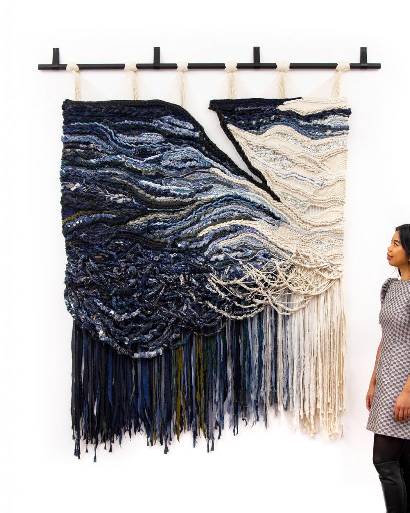 Shades of deep and stormy blue clash into a beige off-white in a large wall hanging overshadowing the woman standing next to it.