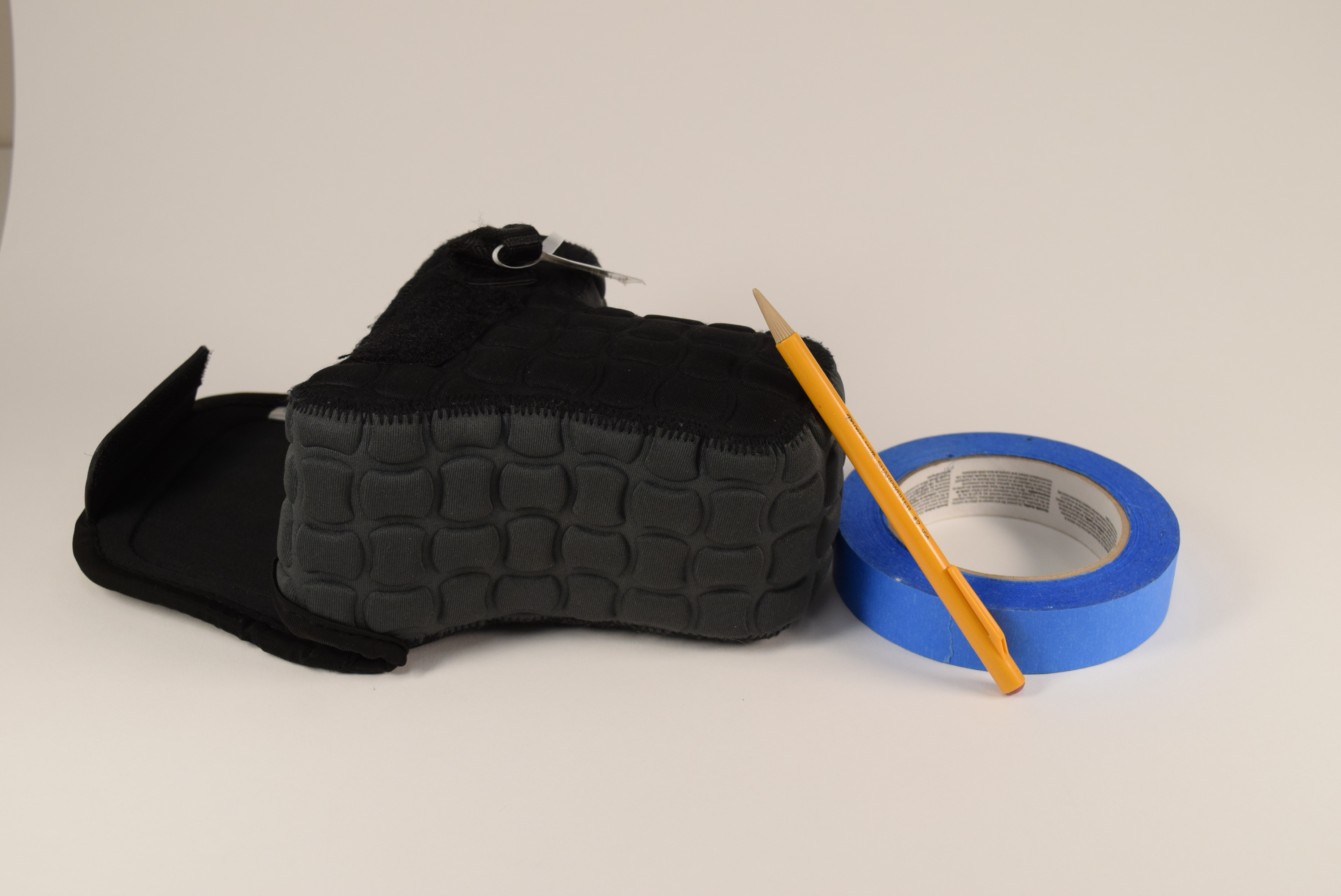 Still life of a camera case, pencil, and roll of blue tape, on a smooth white background.