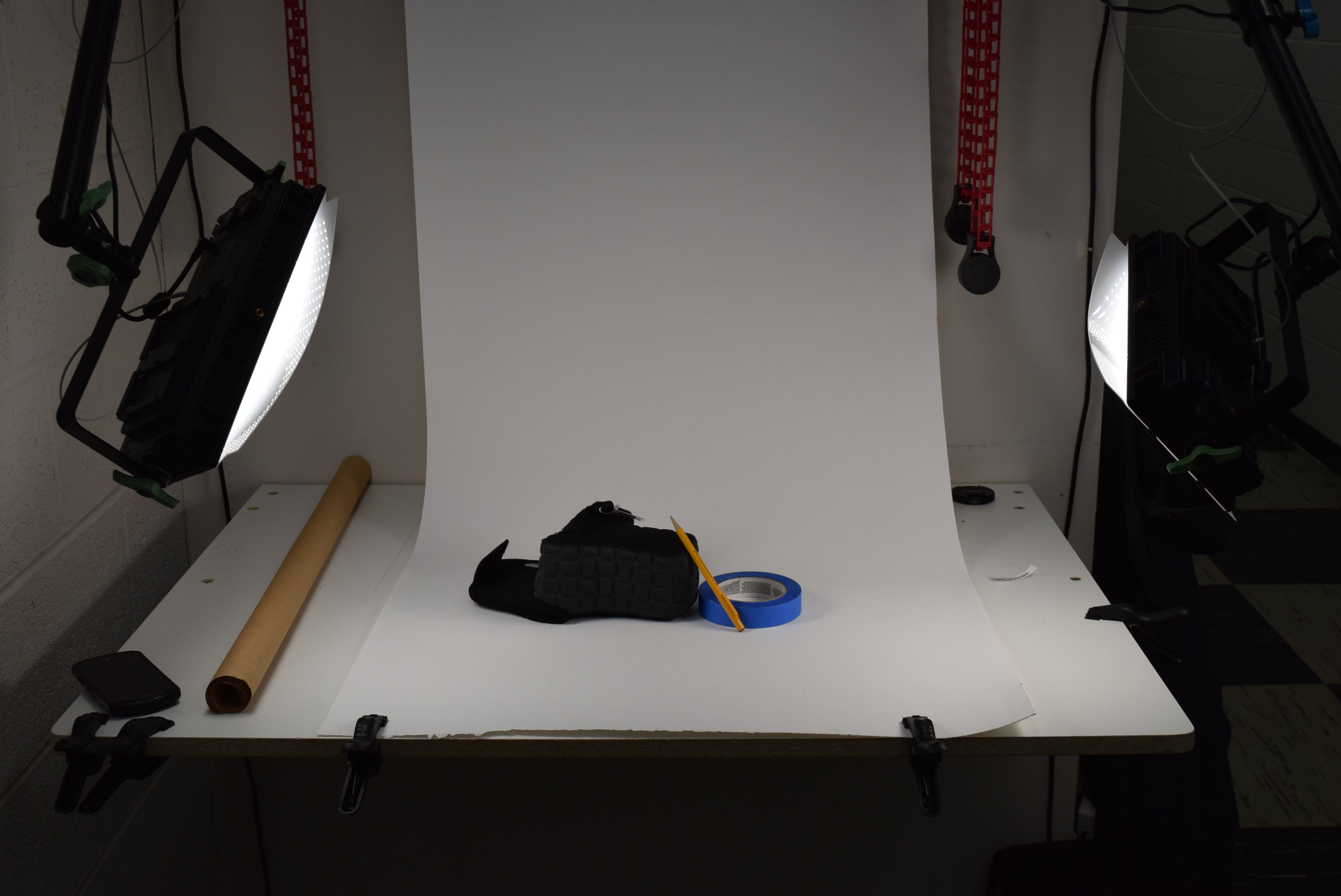 Image of a white table projecting out from the wall, with a roll of slack white paper leading from its front edge back up and out of the frame. Two lights on either side of the table, facing it, light the subject on the paper, which is a small still life of a pencil, camera case, and roll of blue tape.