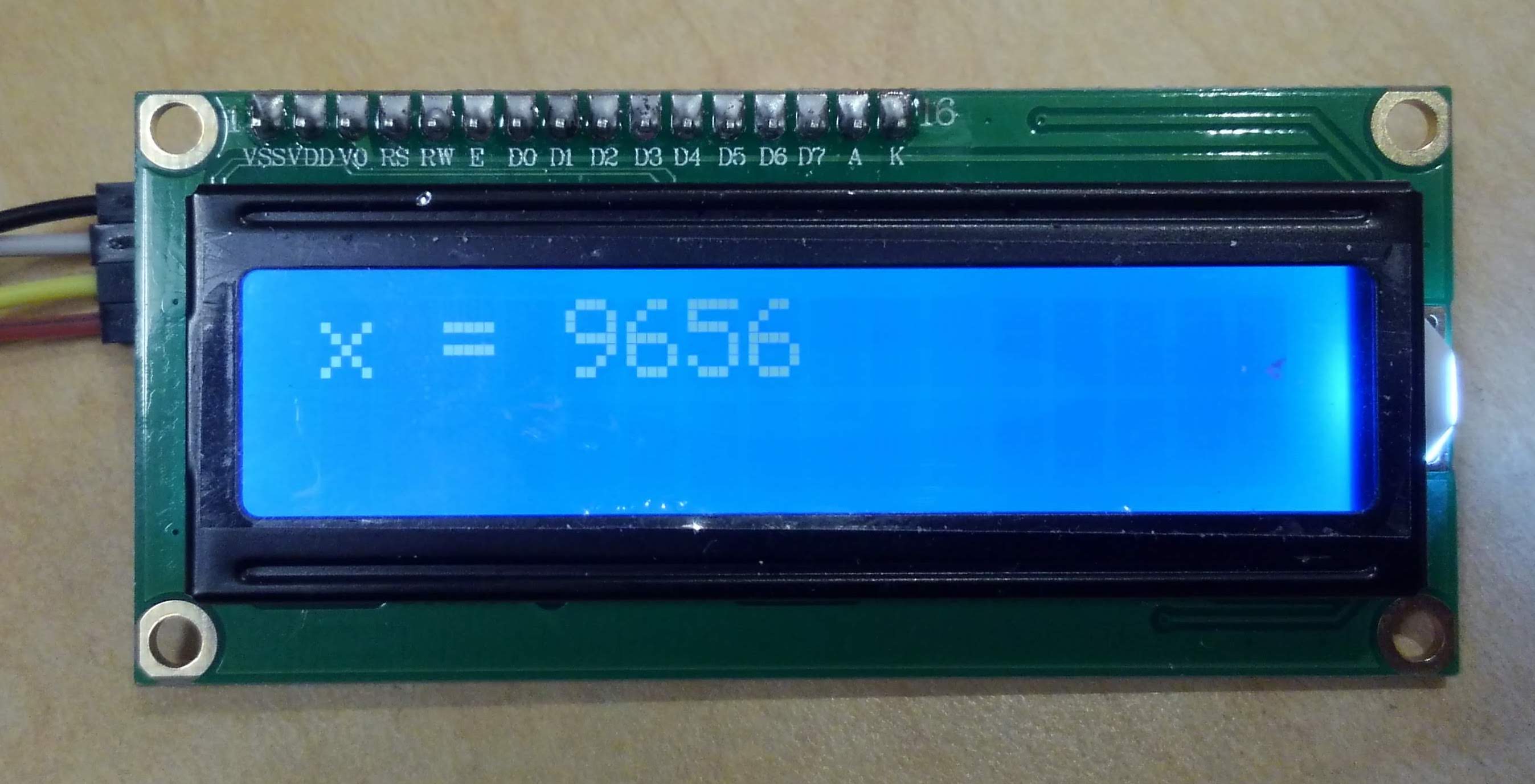 Image of an I2C LCD screen with a blue background, displaying the text "x = 9656" in the upper left corner. Four wires come from the back of the device and run out of frame.
